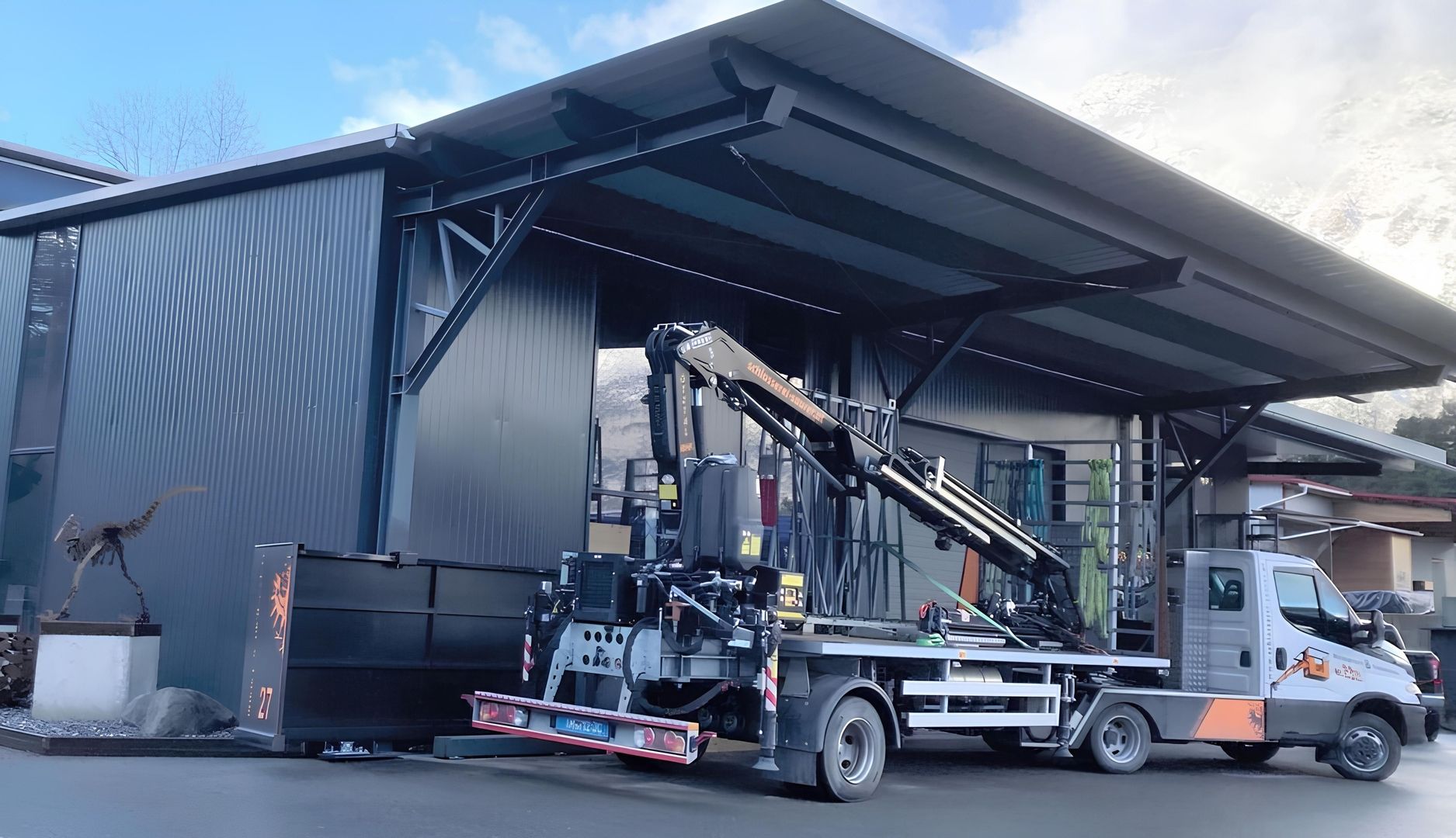 Kleiner Lkw mit Kran vor einem modernen, überdachten Gebäude mit dunkler Fassade und großem Vordach